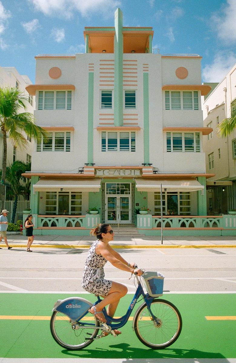 A woman rides a blue bicycle on a green bike lane in front of a white and mint green art deco building.