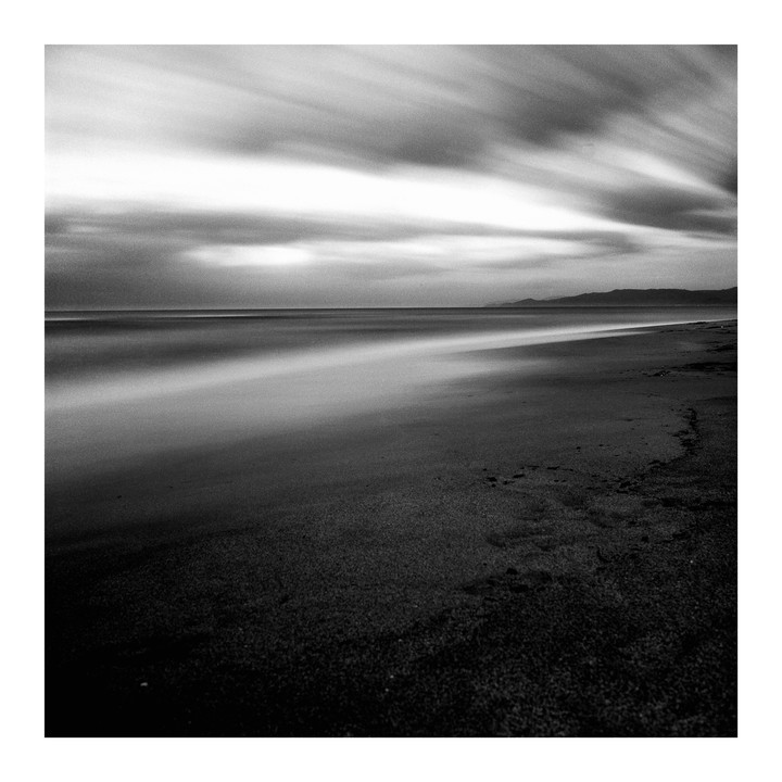 A long exposure captures the movement of clouds and water on a beach with mountains in the distance.