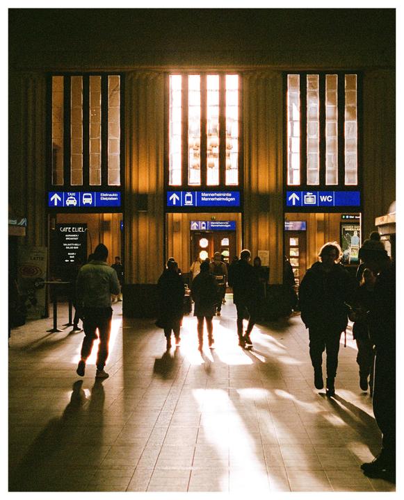 People walk through a train station with light streaming through tall windows and casting long shadows on the floor.