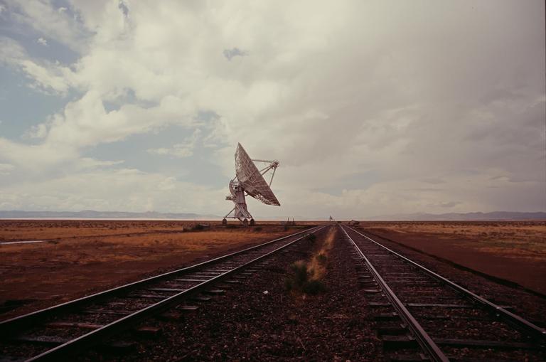A large satellite dish stands next to railroad tracks in a vast, arid landscape under a cloudy sky.