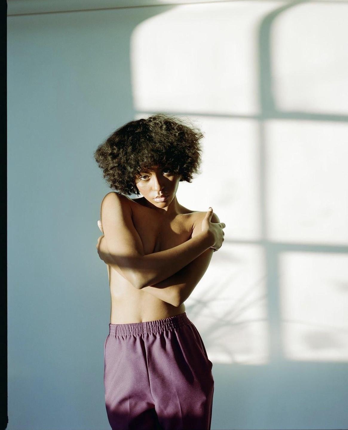 A woman with curly hair stands topless with her arms crossed, wearing purple pants, with shadows on the wall behind her.