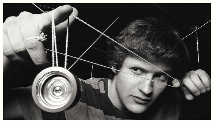 A man with a serious expression looks at the viewer while holding a yo-yo with strings crisscrossing in front of his face.