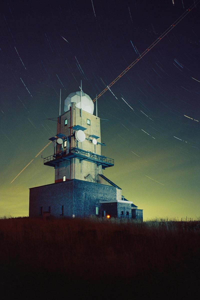 A tall building with a radar dome on top stands under a night sky filled with star trails and a light streak.