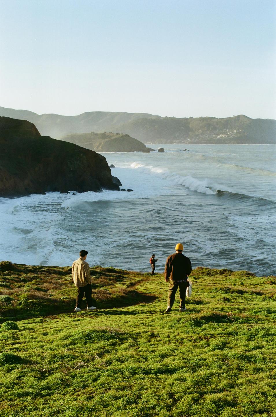 Three men stand on a grassy cliff overlooking a wavy ocean with distant hills under a clear sky.