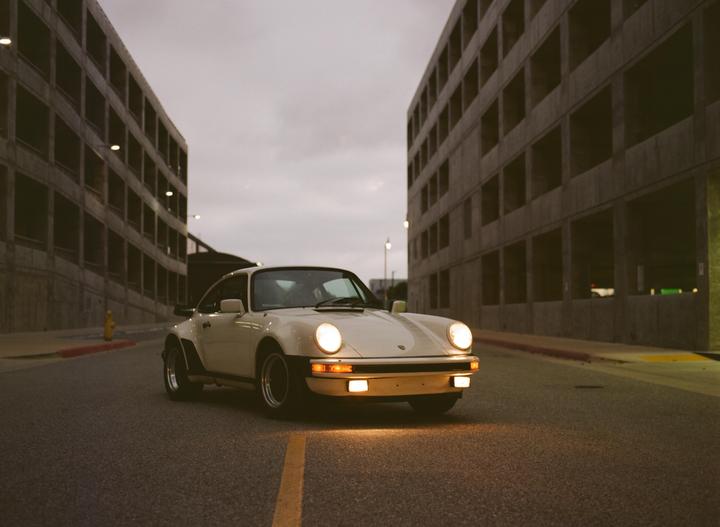 A white Porsche with its headlights on is parked on a street between two large concrete buildings.