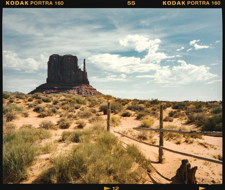 A large rock formation rises from a desert landscape with a wooden fence in the foreground.