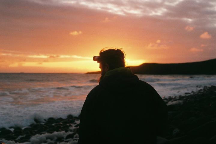 A man with a headlamp stands on a rocky beach looking out at the ocean during sunset.