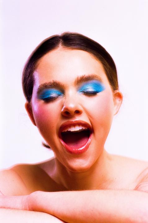 A woman with bright blue eyeshadow and pink blush has her mouth wide open and eyes closed.