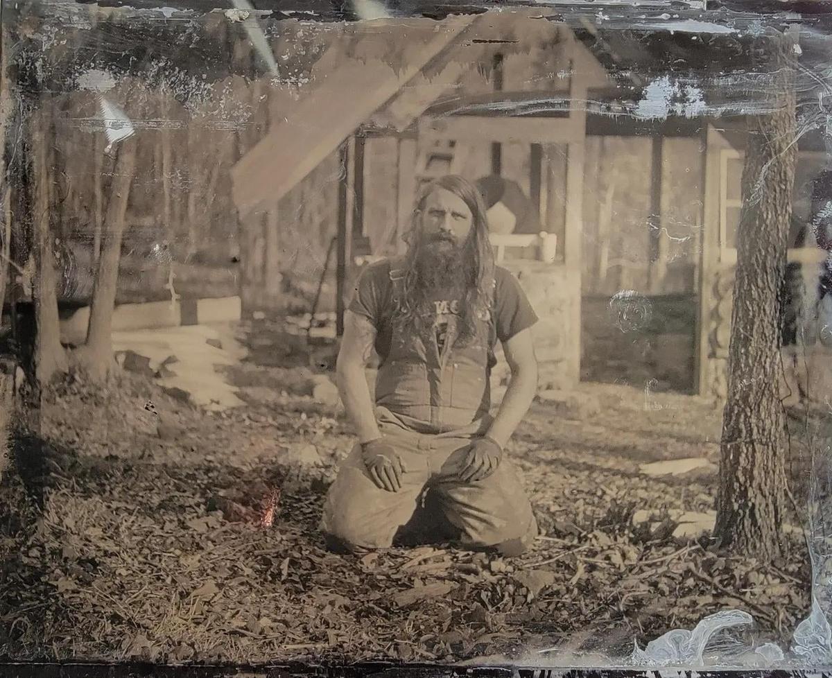 A man with a long beard and hair kneels on the ground in front of a wooden structure.