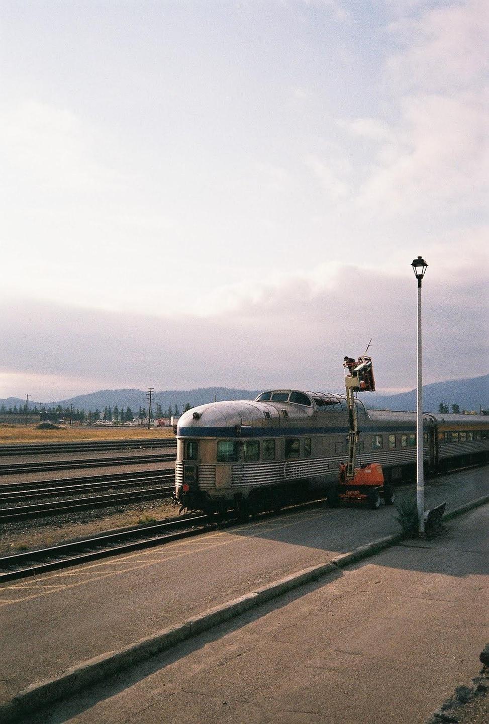 A train is parked on tracks next to a road with a person in a lift working on the train's roof.