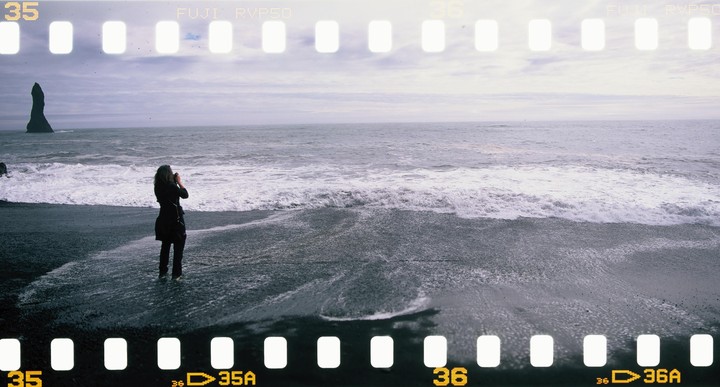 A woman stands on a black sand beach with waves crashing around her feet, facing a sea stack in the distance.