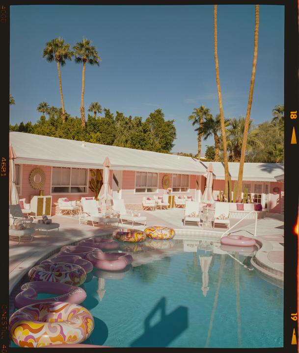 A swimming pool with several inflatable rings floats in front of a pink building with palm trees under a clear blue sky.