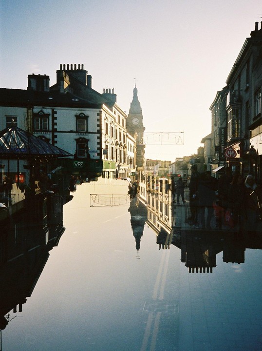 A street scene with buildings and a clock tower is reflected in a large puddle, creating a double exposure effect.