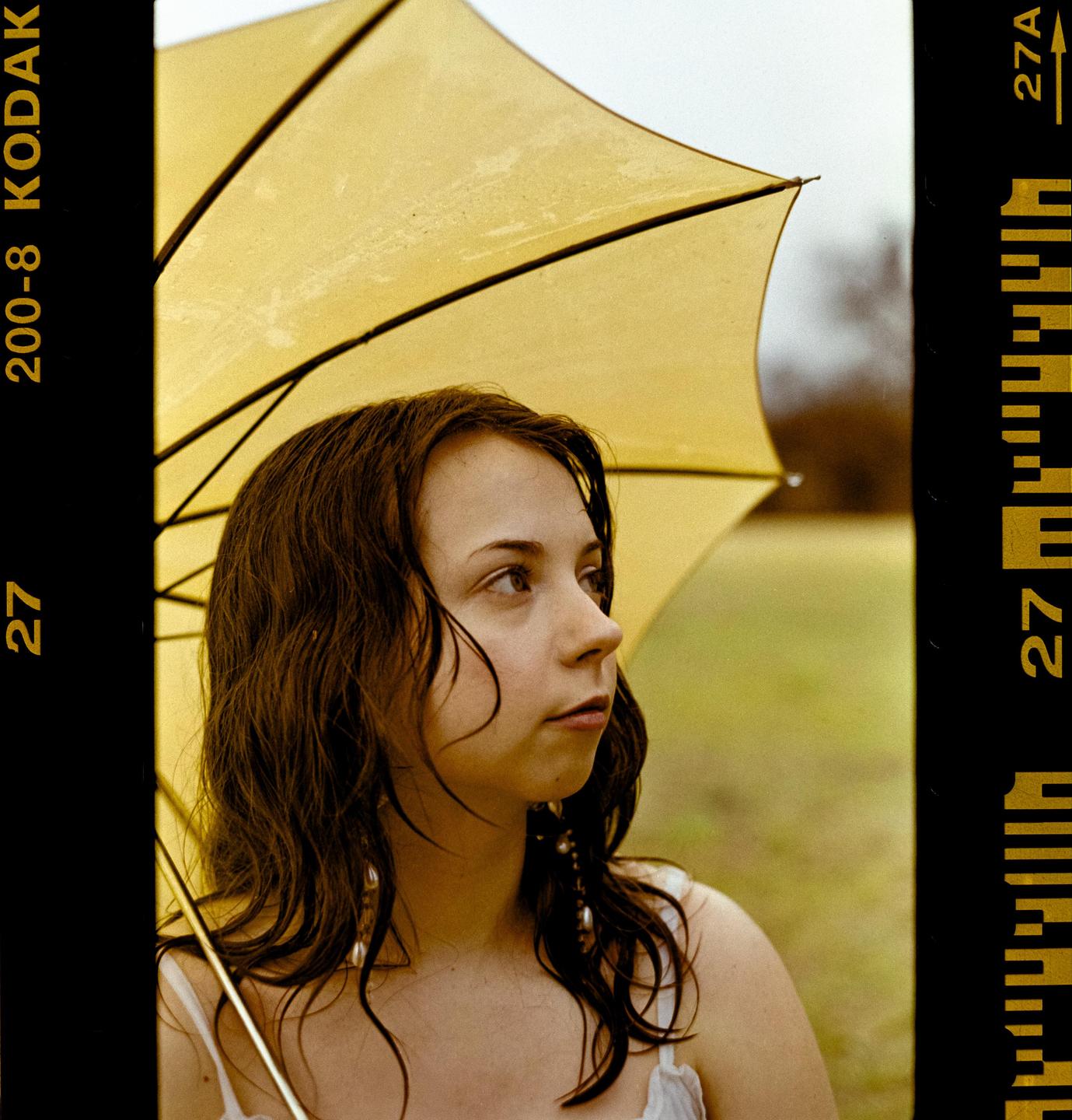 A woman with wet hair stands under a yellow umbrella in a field.