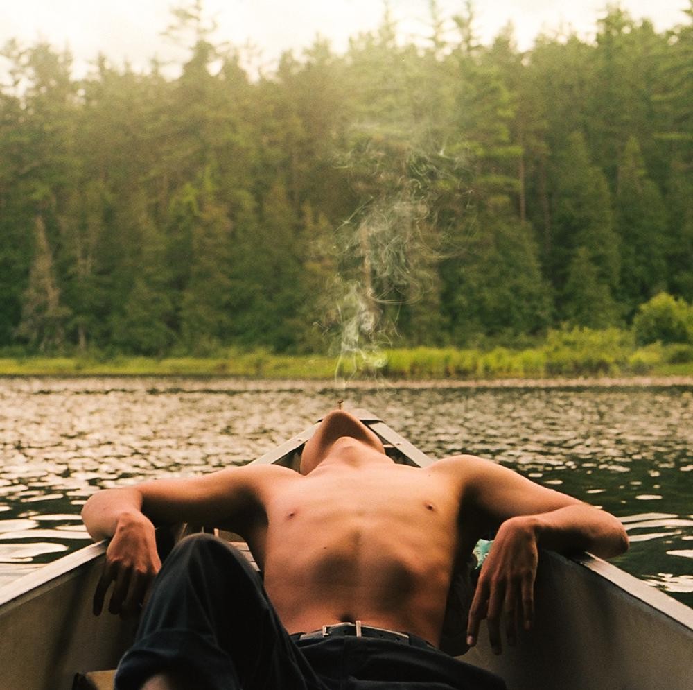 A shirtless man reclines in a canoe on a lake, exhaling smoke towards the sky.