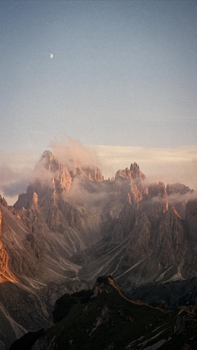 A moon is visible in the sky above a mountain range with clouds and golden hour light.