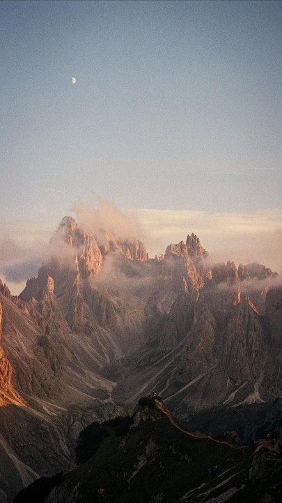 A moon is visible in the sky above a mountain range with clouds and golden hour light.