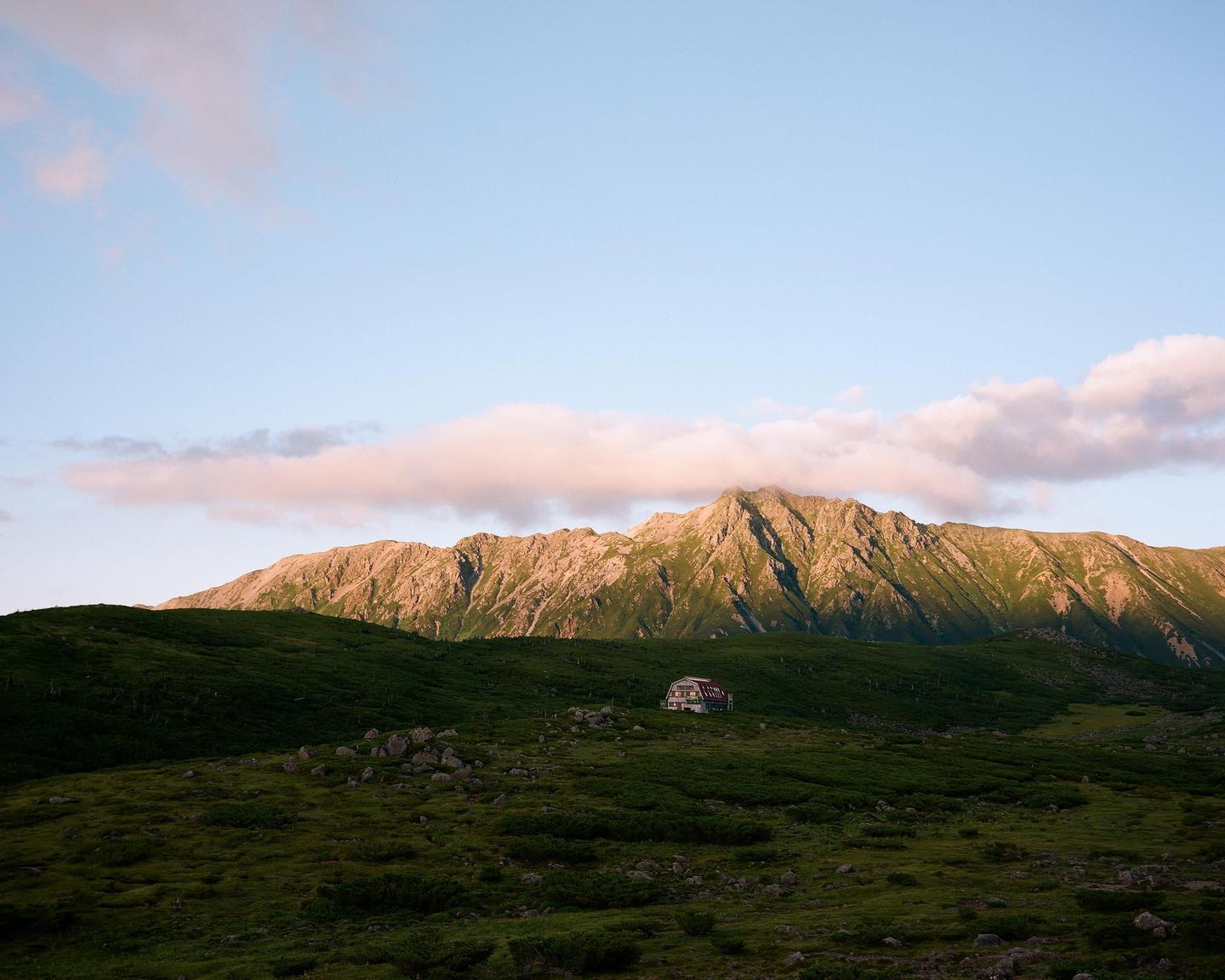 A small building with a red roof sits in a green valley below a large mountain range under a blue sky with pink clouds.