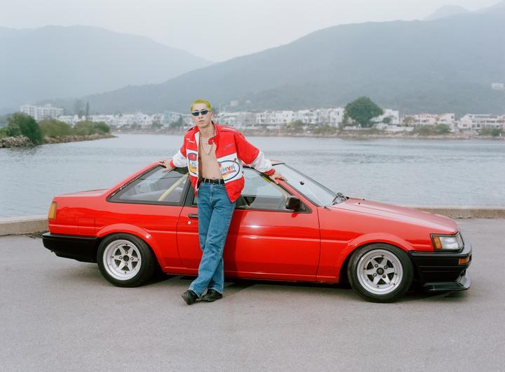 A man with yellow hair and sunglasses leans against a red car by the water with mountains in the background.