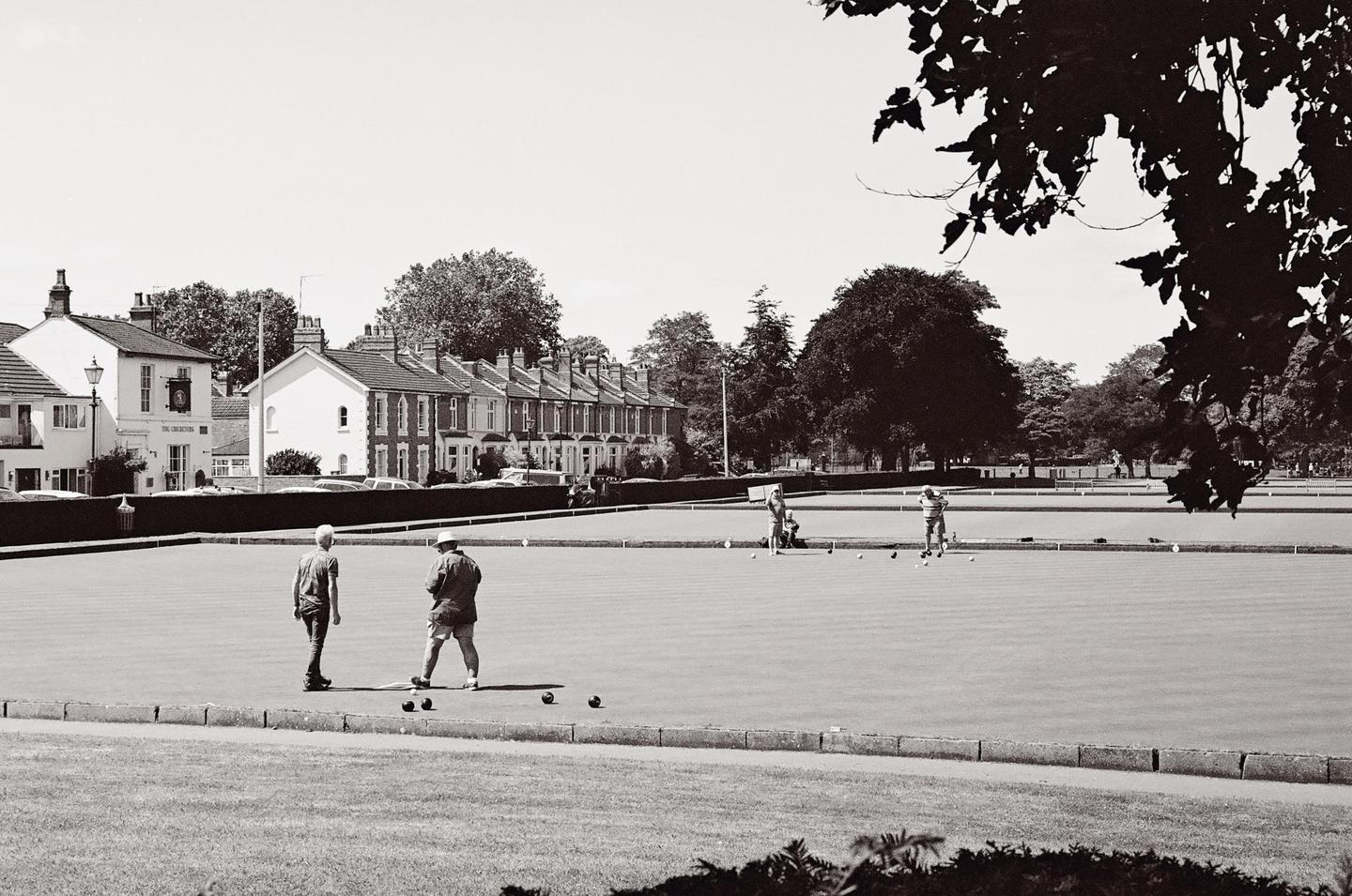 Several people are playing lawn bowls on a green field with houses and trees in the background.