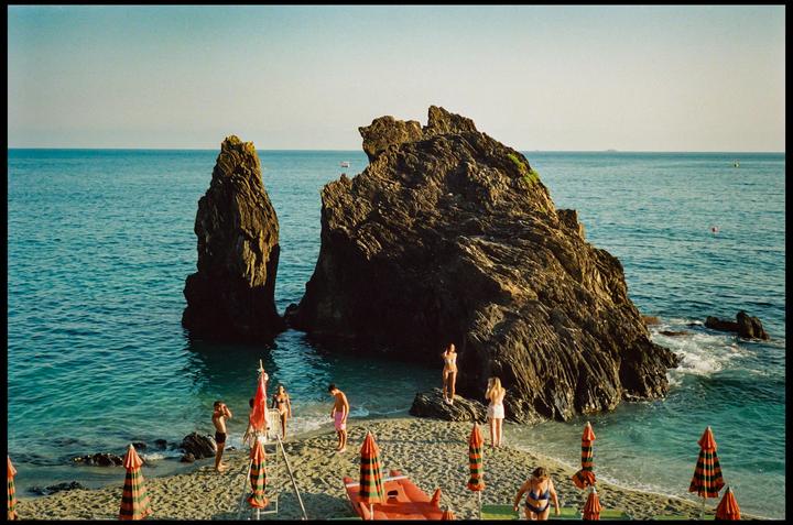 A group of people are on a beach with large rock formations in the water and striped umbrellas on the sand.