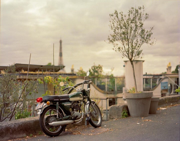 A green motorcycle is parked on a paved area with a tree in a large pot and a houseboat in the background, with the Eiffel Tower visible in the distance.