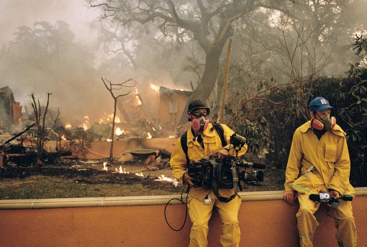 Two men in yellow suits and respirators stand in front of a burning building with smoke and flames.