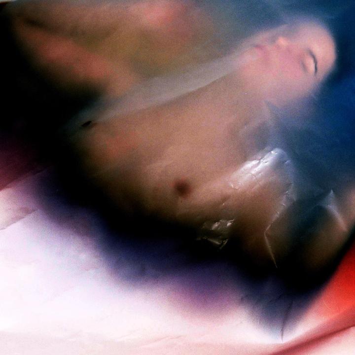 A woman's torso and face are visible through a translucent, wrinkled material with color shifts.