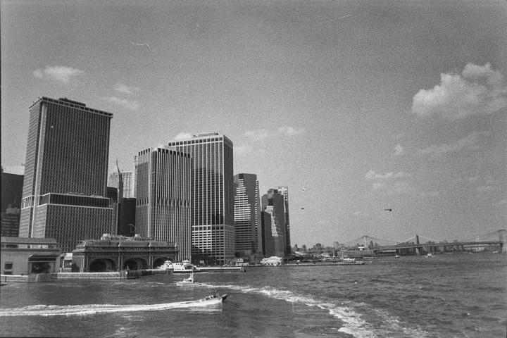 A boat leaves a white wake on the water in front of a city skyline with a bridge in the distance.