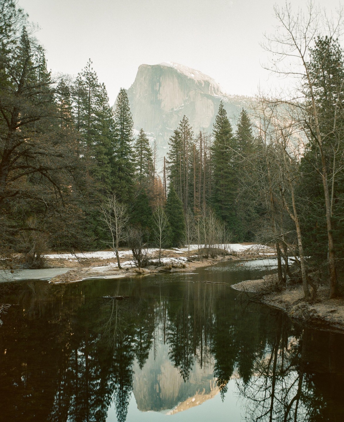A river reflects a mountain and trees under a hazy sky.