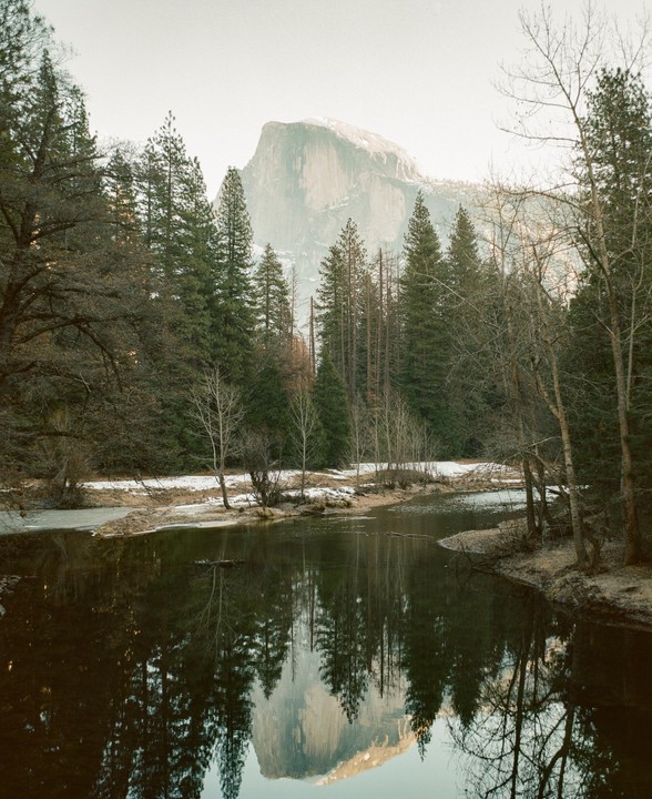 A river reflects a mountain and trees under a hazy sky.