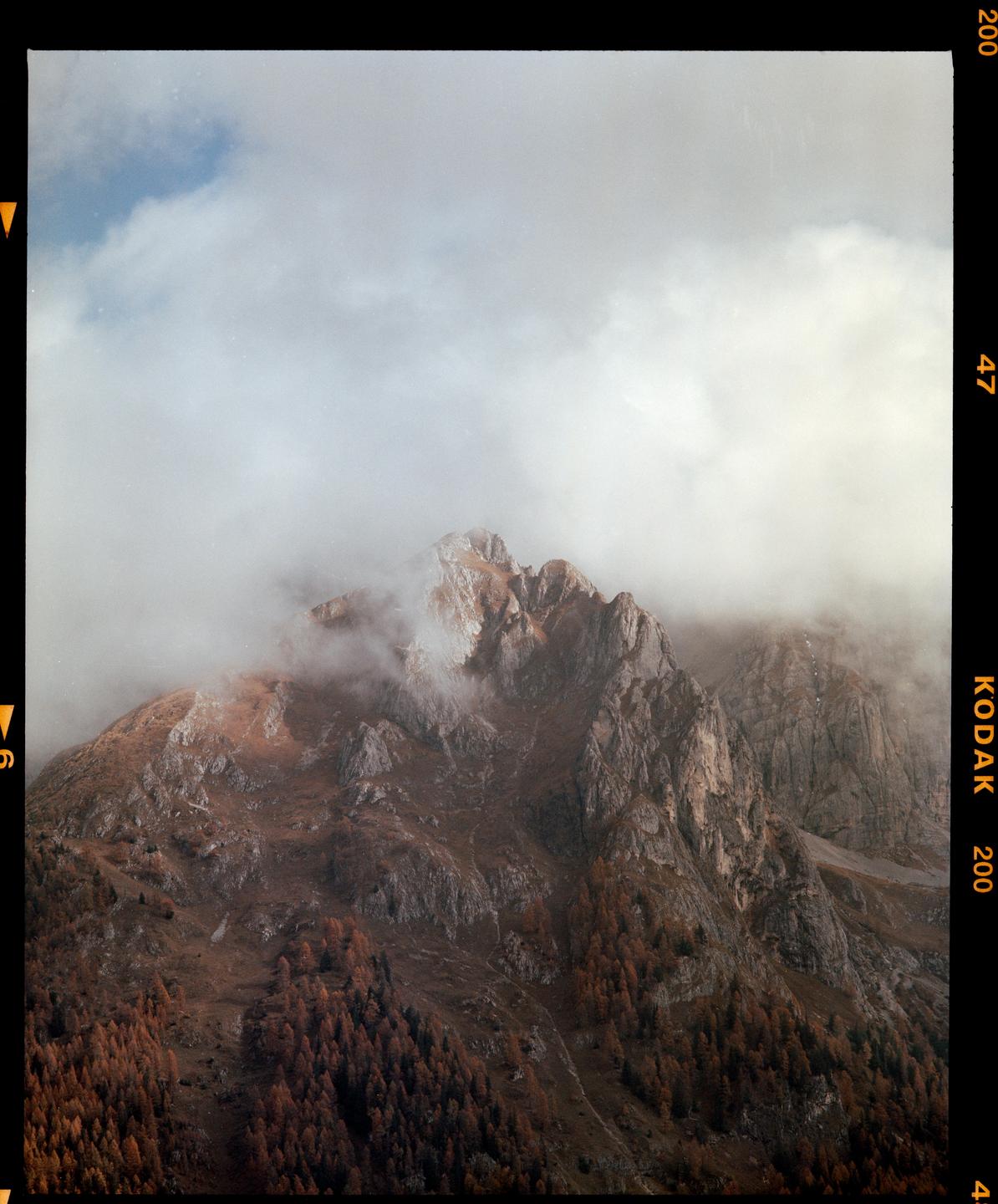 Clouds partially obscure the peaks of a mountain range covered in autumn foliage.