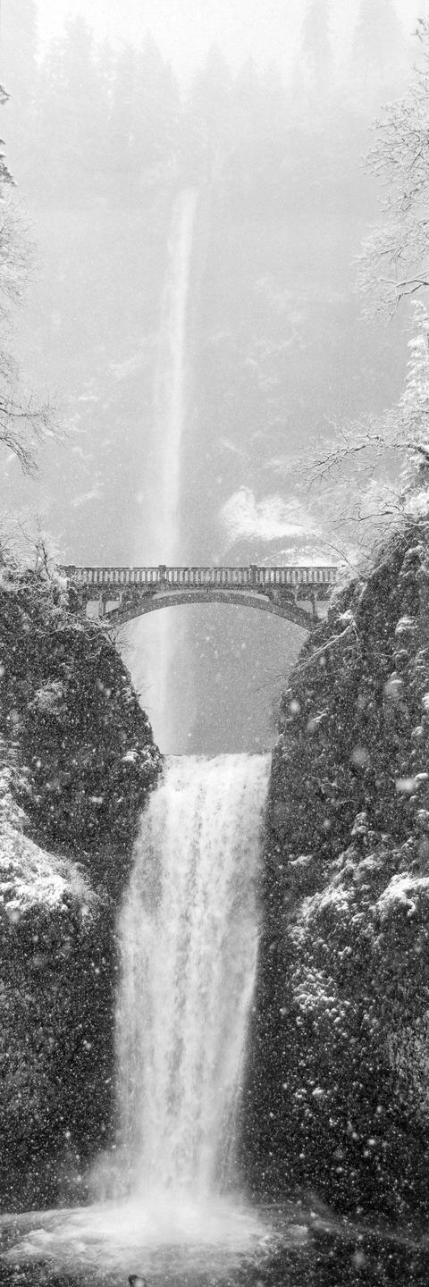 A tall waterfall cascades over a cliff with a bridge spanning the middle section, all covered in snow.