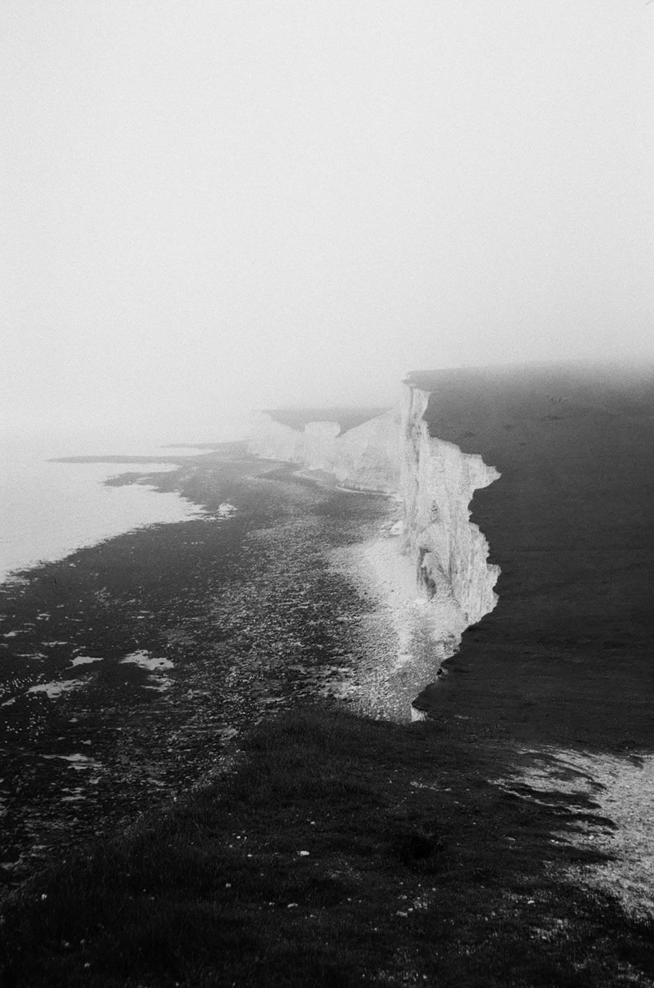 A dramatic black and white landscape shows a long line of white cliffs along a foggy coast.