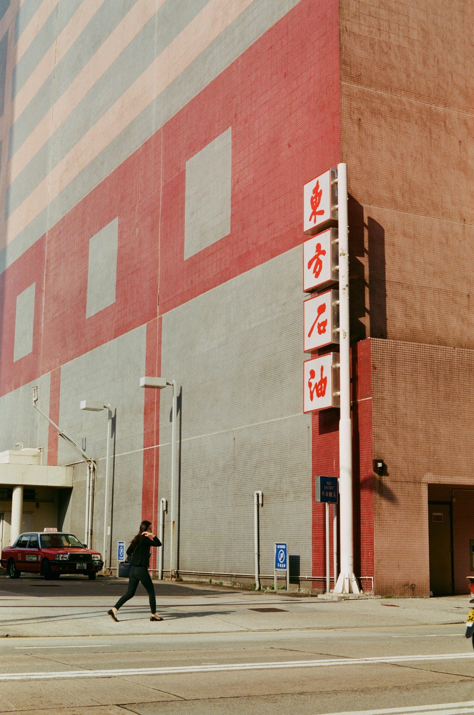 A woman crosses a street in front of a large building with red and gray stripes and a red taxi is parked nearby.