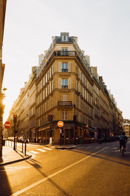 A street corner in Paris is illuminated by the golden hour sun, highlighting a building with many windows and a cafe on the ground floor.