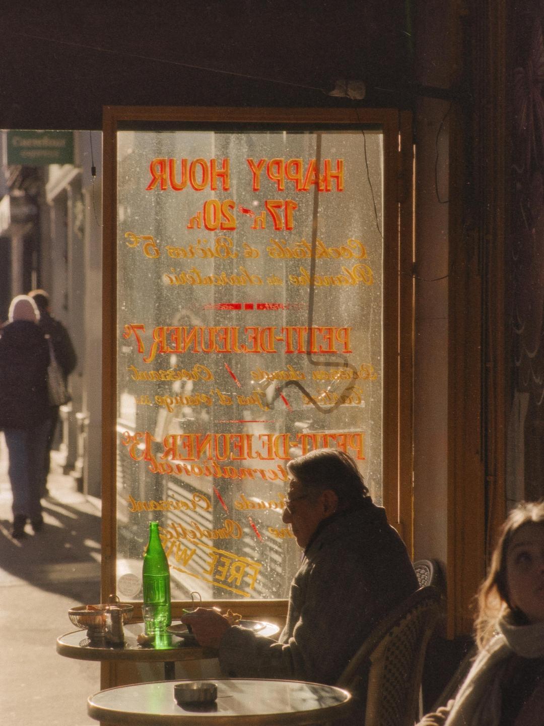 A man sits at a cafe table with a green bottle in front of a window with reversed text.