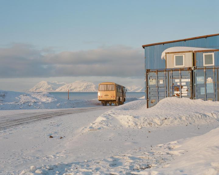 A yellow bus drives on a snowy road next to a blue building made of shipping containers, with mountains and water in the background.