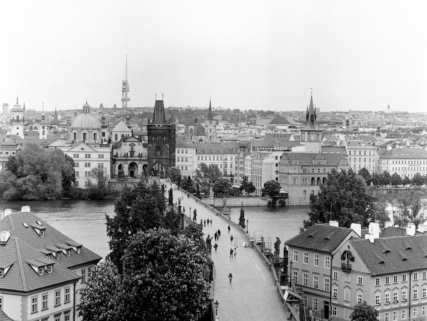 An elevated view shows a bridge with people walking across it, leading to a city with many buildings and a river.