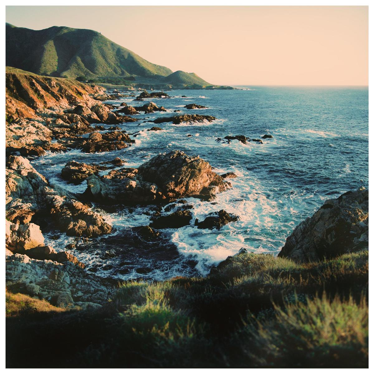 Waves crash against a rocky coastline with mountains in the background.