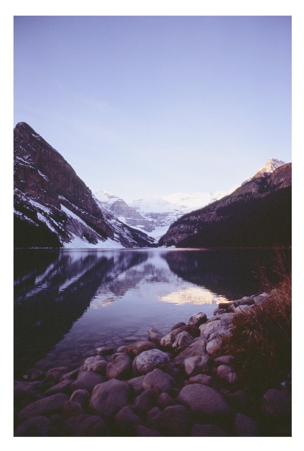 A lake reflects snow-capped mountains under a clear sky before sunrise.