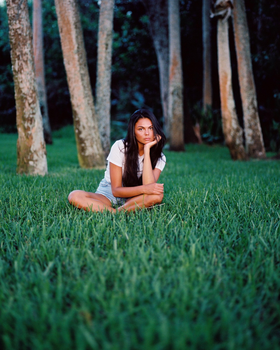 A woman sits cross-legged in a grassy backyard with trees in the background.
