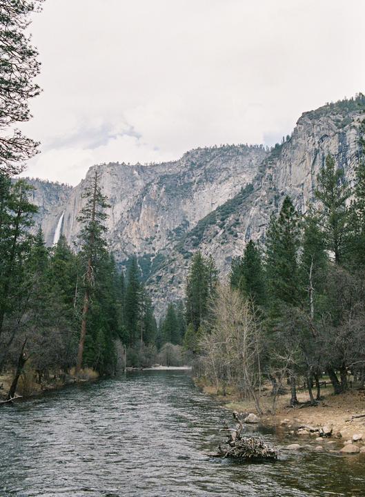 A river flows through a valley surrounded by mountains and trees, with a waterfall visible in the distance.