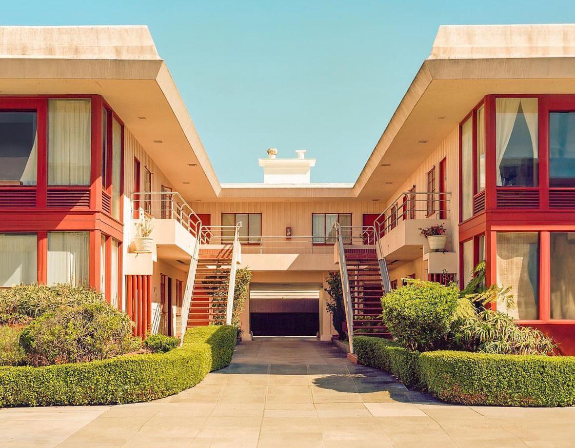 A mid-century modern building with red accents and green hedges is shown under a clear sky.
