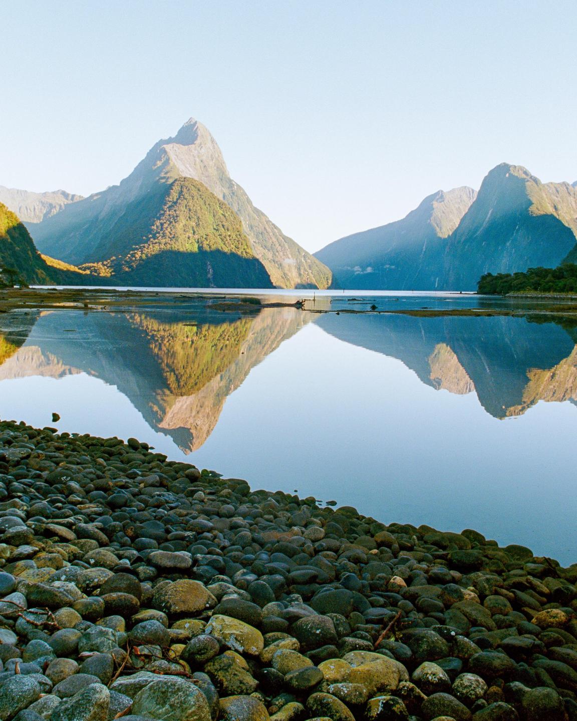 A body of water reflects mountains under a clear sky, with a rocky shore in the foreground.