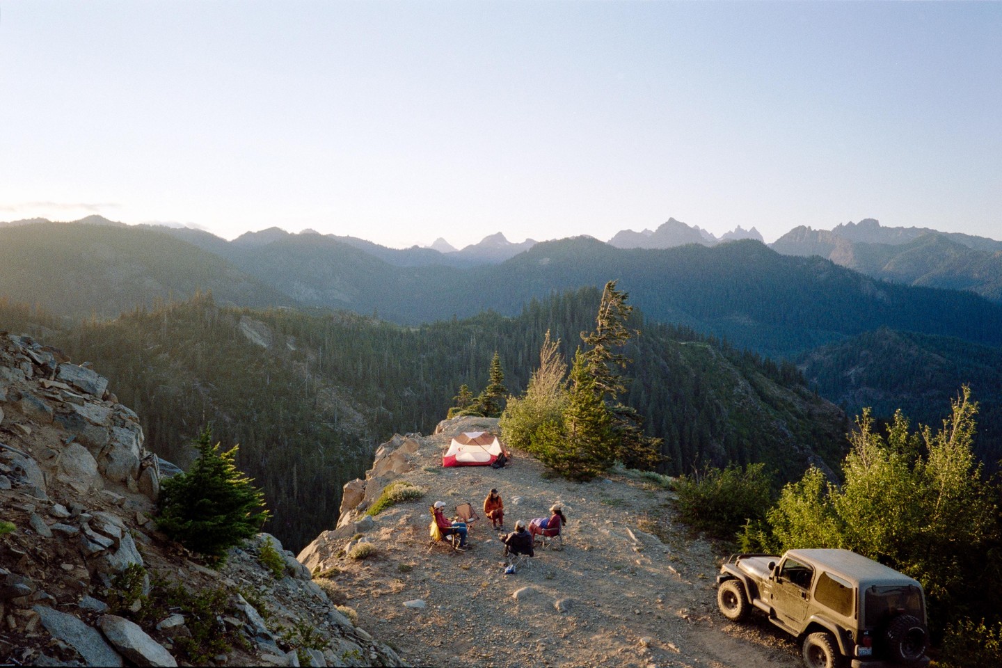 A group of people are camping on a mountain ridge with a tent and a jeep, overlooking a vast forest and mountain range.