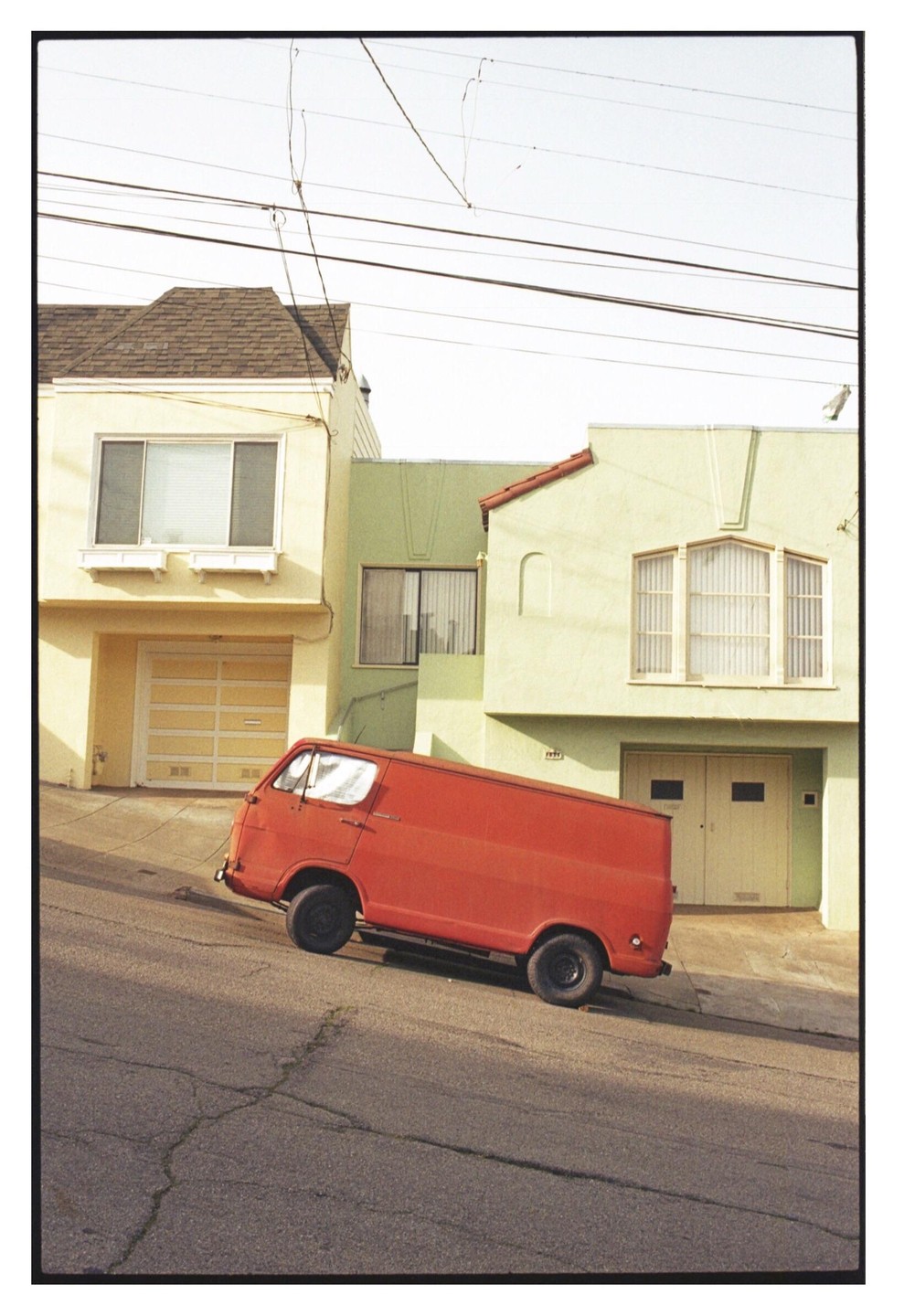 A red van is parked on a steep street in front of two houses with power lines overhead.