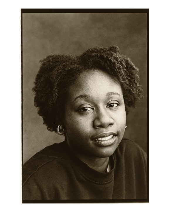 A woman with curly hair and a nose ring looks towards the viewer.