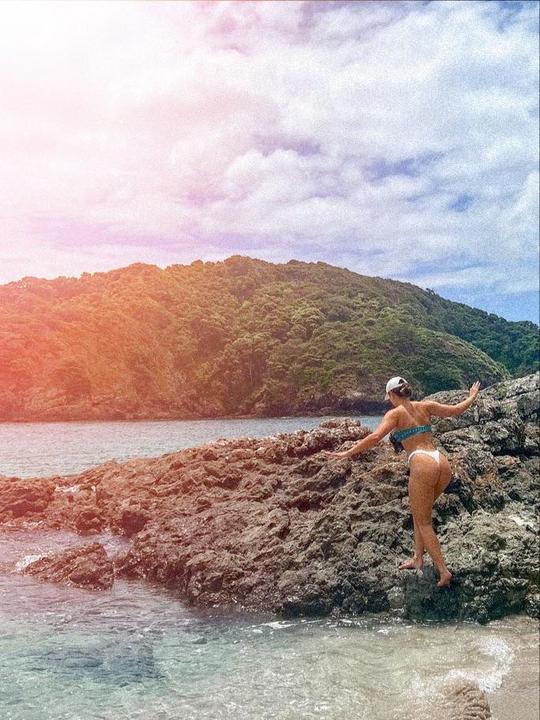 A woman in a bikini climbs over rocks at the edge of a body of water with a forested hill in the background.