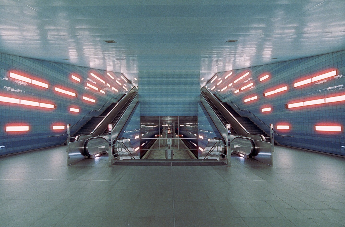 Two escalators lead down to a subway platform with blue tiled walls and orange lights.